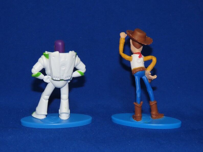 May include: Back view of two plastic action figures, Buzz Lightyear and Woody from the Toy Story franchise. Buzz Lightyear is white with green accents and a purple head. Woody is wearing a brown cowboy hat, a white shirt, a brown belt, blue jeans, and brown boots. Both figures are standing on blue bases.