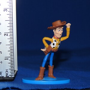 May include: A plastic toy figurine of Woody from the Toy Story franchise. Woody is wearing his signature brown cowboy hat, blue jeans, and a yellow vest with a red bandana. He is holding his right hand up to his hat, and his left hand is on his hip. The figurine is standing on a blue base.