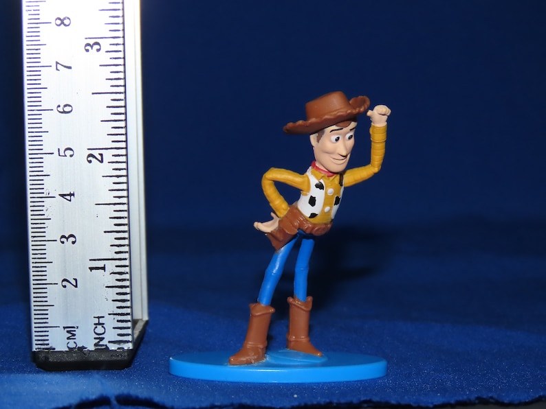 May include: A plastic toy figurine of Woody from the Toy Story franchise. Woody is wearing his signature brown cowboy hat, blue jeans, and a yellow vest with a red bandana. He is holding his right hand up to his hat, and his left hand is on his hip. The figurine is standing on a blue base.