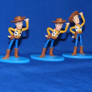 May include: Three plastic toy figures of Woody from Toy Story. Each figure is wearing a brown cowboy hat, a yellow shirt with a red bandana, blue jeans, and brown boots. The figures are standing on blue bases.