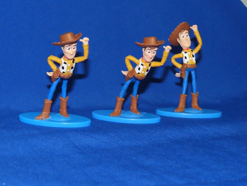 May include: Three plastic toy figures of Woody from Toy Story. Each figure is wearing a brown cowboy hat, a yellow shirt with a red bandana, blue jeans, and brown boots. The figures are standing on blue bases.