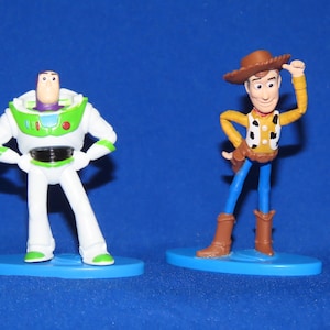 May include: Toy Story characters Woody and Buzz Lightyear figurines. Woody is wearing a brown cowboy hat, a yellow vest with a red bandana, and blue jeans. Buzz Lightyear is wearing a white and green spacesuit with a purple helmet. Both figurines are standing on blue bases.