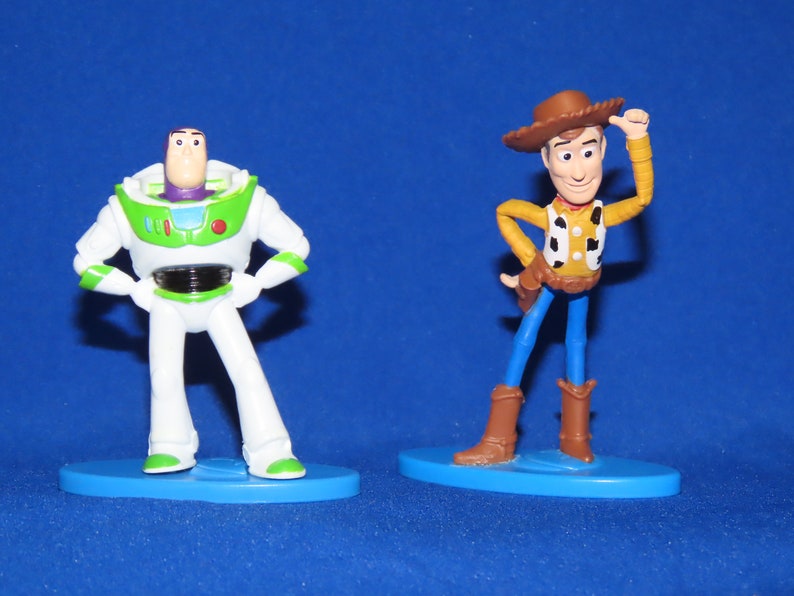 May include: Toy Story characters Woody and Buzz Lightyear figurines. Woody is wearing a brown cowboy hat, a yellow vest with a red bandana, and blue jeans. Buzz Lightyear is wearing a white and green spacesuit with a purple helmet. Both figurines are standing on blue bases.