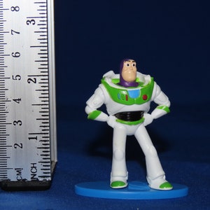 May include: A white and green plastic toy figurine of Buzz Lightyear from the Toy Story franchise. The figurine is standing on a blue base and is approximately 5 cm tall.