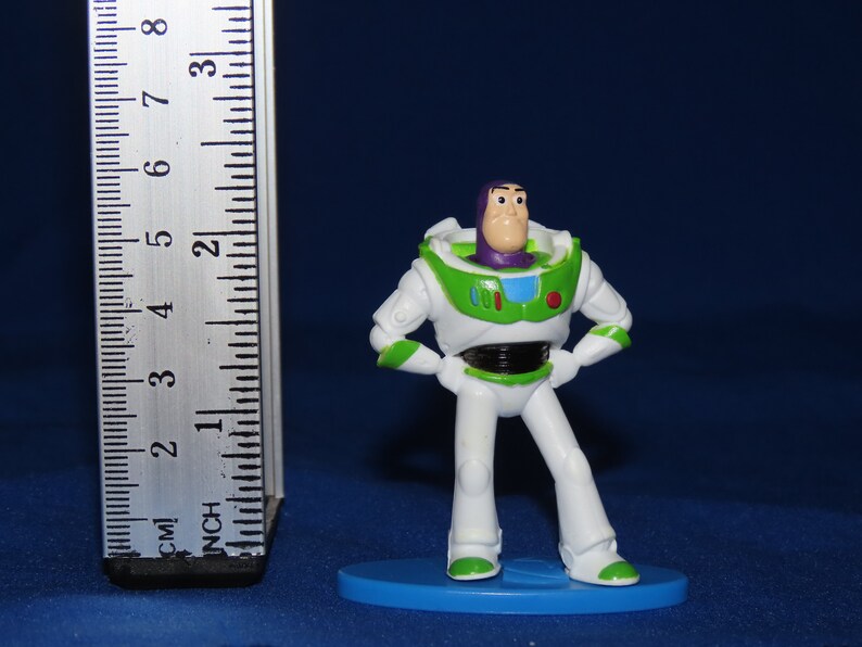 May include: A white and green plastic toy figurine of Buzz Lightyear from the Toy Story franchise. The figurine is standing on a blue base and is approximately 5 cm tall.