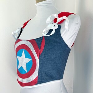K&ouml;nnte beinhalten: Ein bauchfreies Top im Jeans-Stil mit einem Captain America Schild-Design. Es hat rote Schultertr&auml;ger und wei&szlig;e Schleifenb&auml;nder. Das Schild-Design zeigt einen wei&szlig;en Stern auf einem blauen Kreis, umgeben von roten und wei&szlig;en Streifen.