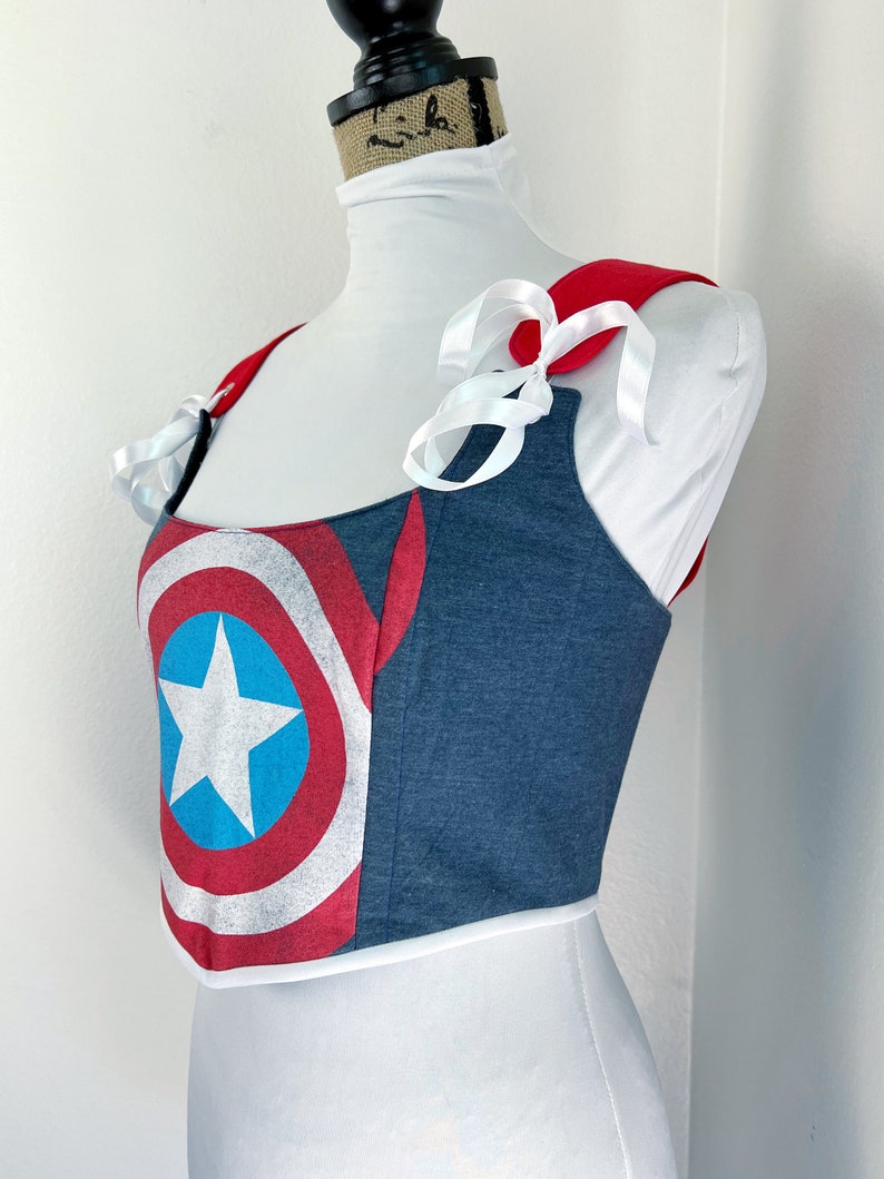 K&ouml;nnte beinhalten: Ein bauchfreies Top im Jeans-Stil mit einem Captain America Schild-Design. Es hat rote Schultertr&auml;ger und wei&szlig;e Schleifenb&auml;nder. Das Schild-Design zeigt einen wei&szlig;en Stern auf einem blauen Kreis, umgeben von roten und wei&szlig;en Streifen.