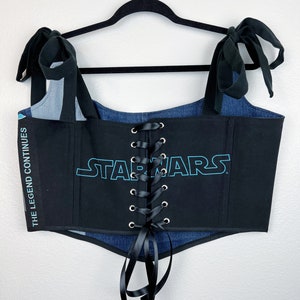K&ouml;nnte beinhalten: Ein schwarzes Korsett-Top mit dem Star Wars-Logo in T&uuml;rkis. Das Oberteil hat Jeans-Akzente und schwarze B&auml;nder zum Schn&uuml;ren. Die Schultertr&auml;ger sind schwarz und zu Schleifen gebunden. Der Text "THE LEGEND CONTINUES" ist links vertikal aufgedruckt.