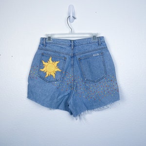 May include: Light blue denim shorts with a yellow sun embroidered on the back pocket and a pattern of colourful dots on the back. The shorts have a frayed hem.