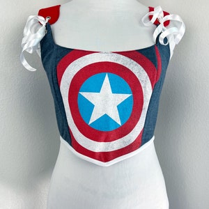 K&ouml;nnte beinhalten: Ein Corsagen-Top im Jeans-Stil mit dem Captain America-Schild-Design in Rot, Wei&szlig; und Blau. Es hat wei&szlig;e Schleifenb&auml;nder an den Schultern und eine wei&szlig;e Bord&uuml;re. Die Corsage wird auf einer Schaufensterpuppe pr&auml;sentiert.