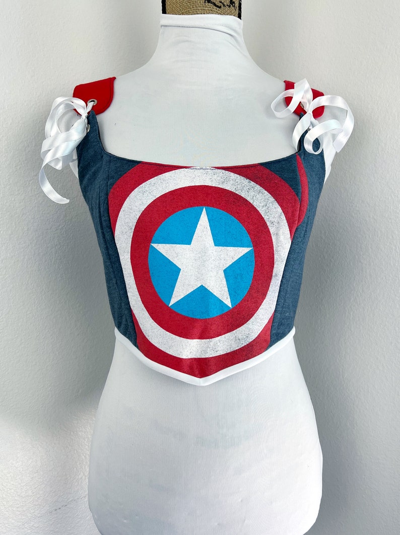 K&ouml;nnte beinhalten: Ein Corsagen-Top im Jeans-Stil mit dem Captain America-Schild-Design in Rot, Wei&szlig; und Blau. Es hat wei&szlig;e Schleifenb&auml;nder an den Schultern und eine wei&szlig;e Bord&uuml;re. Die Corsage wird auf einer Schaufensterpuppe pr&auml;sentiert.