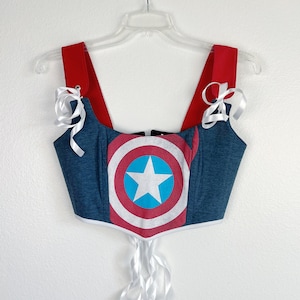 K&ouml;nnte beinhalten: Ein blau-rotes Korsett-Top mit dem Design des Captain America-Schildes. Das Top hat rote Tr&auml;ger und wei&szlig;e Schleifenb&auml;nder. Die Schildgrafik ist mittig auf der Vorderseite angebracht, mit einem wei&szlig;en Stern auf einem blauen Kreis.
