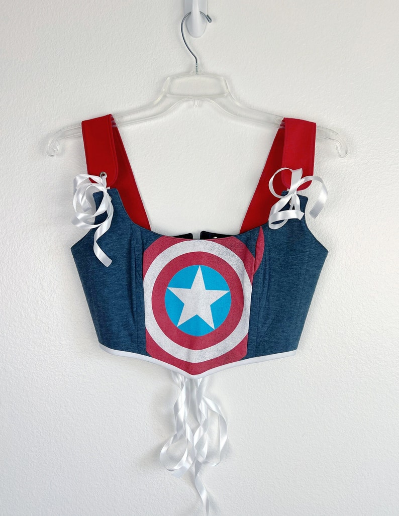 K&ouml;nnte beinhalten: Ein blau-rotes Korsett-Top mit dem Design des Captain America-Schildes. Das Top hat rote Tr&auml;ger und wei&szlig;e Schleifenb&auml;nder. Die Schildgrafik ist mittig auf der Vorderseite angebracht, mit einem wei&szlig;en Stern auf einem blauen Kreis.