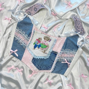 Puede incluir: Top de mezclilla patchwork con un bolsillo, con detalles de encaje y lazos rosas. El top tiene una estética vintage, con un panel central que representa una escena de dibujos animados. Los lazos rosas están esparcidos sobre un fondo de satén blanco.