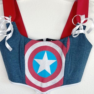K&ouml;nnte beinhalten: Ein blau-rotes Korsett-Top mit dem Captain America-Schild-Design. Das Top hat rote Tr&auml;ger und wei&szlig;e Schleifenb&auml;nder. Das Korsett h&auml;ngt an einem transparenten Kleiderb&uuml;gel vor wei&szlig;em Hintergrund.