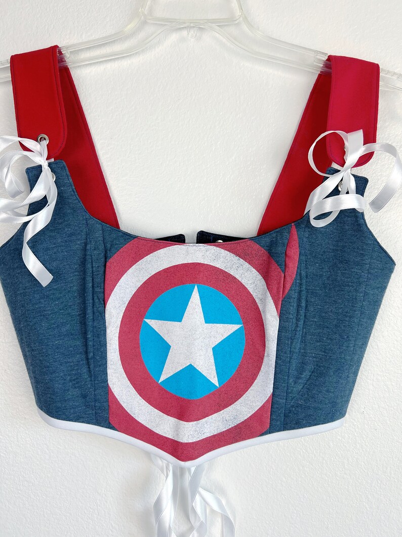 K&ouml;nnte beinhalten: Ein blau-rotes Korsett-Top mit dem Captain America-Schild-Design. Das Top hat rote Tr&auml;ger und wei&szlig;e Schleifenb&auml;nder. Das Korsett h&auml;ngt an einem transparenten Kleiderb&uuml;gel vor wei&szlig;em Hintergrund.
