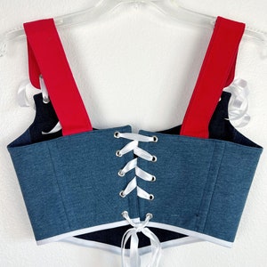 K&ouml;nnte beinhalten: Ein Corsagen-Top im Jeans-Stil mit roten Tr&auml;gern und wei&szlig;er Schn&uuml;rung. Die Corsage hat einen wei&szlig;en Besatz am unteren Rand und wird auf einem transparenten Kleiderb&uuml;gel vor wei&szlig;em Hintergrund pr&auml;sentiert. Das Top ist eine Mischung aus Blau, Rot und Wei&szlig;.