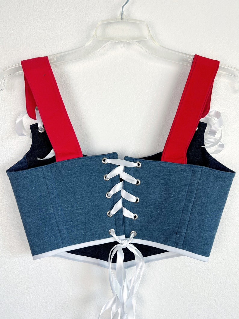 K&ouml;nnte beinhalten: Ein Corsagen-Top im Jeans-Stil mit roten Tr&auml;gern und wei&szlig;er Schn&uuml;rung. Die Corsage hat einen wei&szlig;en Besatz am unteren Rand und wird auf einem transparenten Kleiderb&uuml;gel vor wei&szlig;em Hintergrund pr&auml;sentiert. Das Top ist eine Mischung aus Blau, Rot und Wei&szlig;.