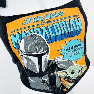 K&ouml;nnte beinhalten: Schwarzes Crop-Top mit einem Grafikdesign aus der Star Wars-Serie "The Mandalorian". Das Design zeigt den Helm des Mandalorianers, Grogu und den Text "This is more than I signed up for". Das Top hat Bindetr&auml;ger.