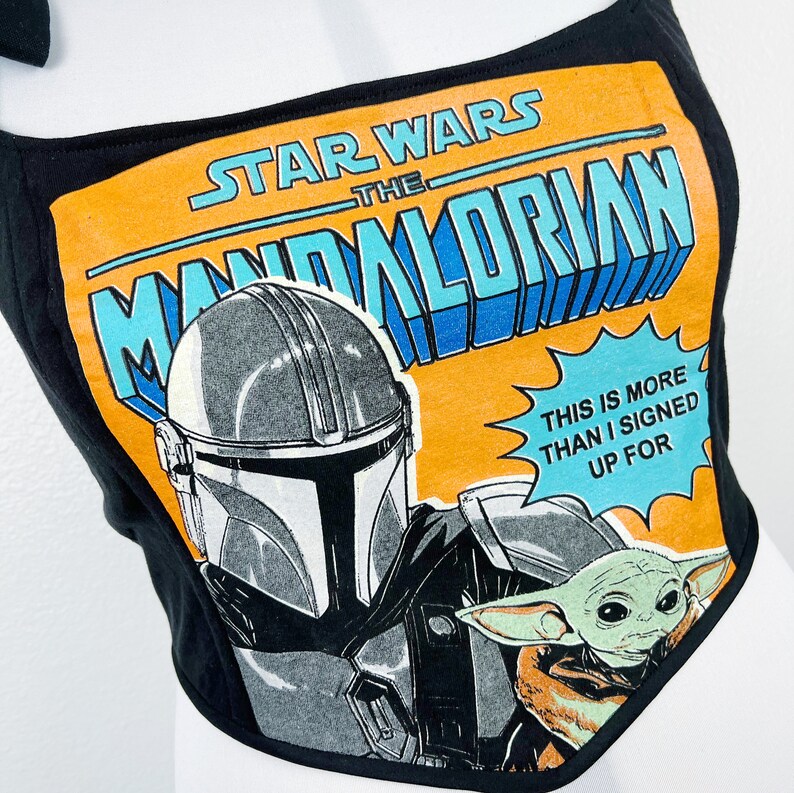 K&ouml;nnte beinhalten: Schwarzes Crop-Top mit einem Grafikdesign aus der Star Wars-Serie "The Mandalorian". Das Design zeigt den Helm des Mandalorianers, Grogu und den Text "This is more than I signed up for". Das Top hat Bindetr&auml;ger.