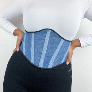 May include: A denim corset with a black trim. The corset features vertical panels of varying shades of blue denim. The garment is worn over a white long-sleeved top and black leggings.