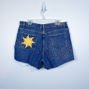 Peut inclure: Short en jean bleu avec un soleil jaune brodé sur la poche arrière et des points colorés sur l'arrière du short.