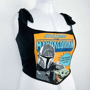 K&ouml;nnte beinhalten: Schwarzes Crop-Top mit Bindetr&auml;gern mit einer Star Wars The Mandalorian Comic-Grafik. Die Grafik zeigt den Mandalorianer, Grogu und den Text "THIS IS MORE THAN I SIGNED UP FOR."
