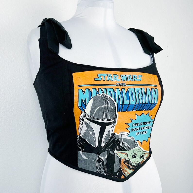 K&ouml;nnte beinhalten: Schwarzes Crop-Top mit Bindetr&auml;gern mit einer Star Wars The Mandalorian Comic-Grafik. Die Grafik zeigt den Mandalorianer, Grogu und den Text "THIS IS MORE THAN I SIGNED UP FOR."