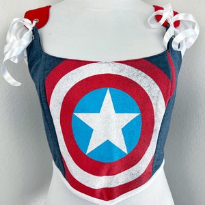 K&ouml;nnte beinhalten: Ein Korsett im Captain America-Schild-Design. Das Oberteil hat ein blaues, rotes und wei&szlig;es Schilddesign mit einem wei&szlig;en Stern auf einem blauen Kreis. Es hat rote Schultergurte mit wei&szlig;en Schleifen. Das Korsett wird auf einer wei&szlig;en Schaufensterpuppe pr&auml;sentiert.