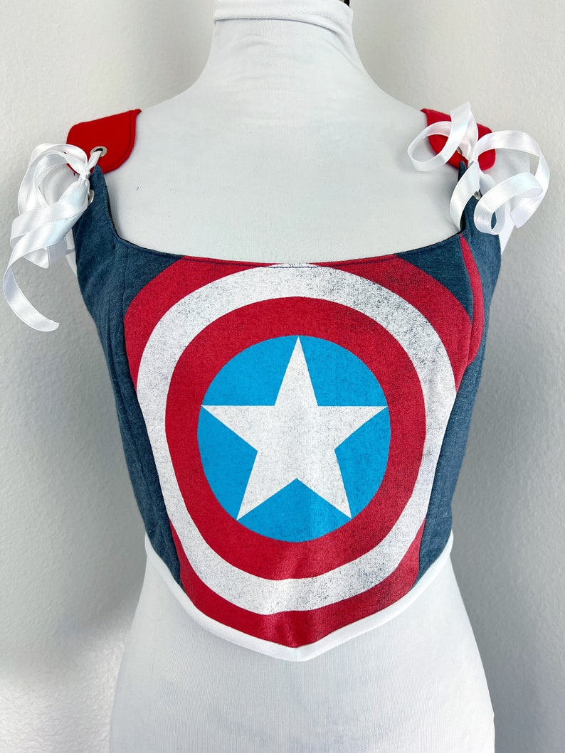 K&ouml;nnte beinhalten: Ein Korsett im Captain America-Schild-Design. Das Oberteil hat ein blaues, rotes und wei&szlig;es Schilddesign mit einem wei&szlig;en Stern auf einem blauen Kreis. Es hat rote Schultergurte mit wei&szlig;en Schleifen. Das Korsett wird auf einer wei&szlig;en Schaufensterpuppe pr&auml;sentiert.
