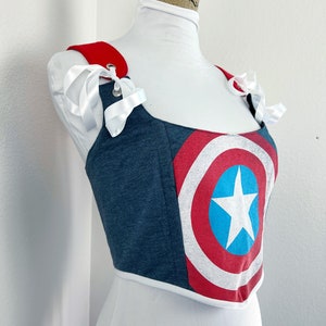 K&ouml;nnte beinhalten: Ein blau-rotes Korsett-Top mit dem Design des Captain America-Schildes. Das Top hat rote Tr&auml;ger mit wei&szlig;en Schleifen und eine wei&szlig;e Bord&uuml;re. Das Korsett wird auf einer wei&szlig;en Schaufensterpuppe pr&auml;sentiert.