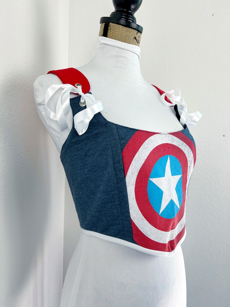K&ouml;nnte beinhalten: Ein blau-rotes Korsett-Top mit dem Design des Captain America-Schildes. Das Top hat rote Tr&auml;ger mit wei&szlig;en Schleifen und eine wei&szlig;e Bord&uuml;re. Das Korsett wird auf einer wei&szlig;en Schaufensterpuppe pr&auml;sentiert.