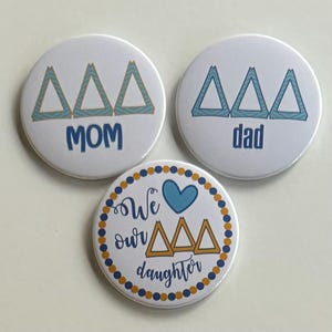 Puede incluir: Tres botones redondos blancos con diseños azules y dorados. El botón superior izquierdo dice "MOM" con tres triángulos azules con detalles dorados. El botón superior derecho dice "dad" con tres triángulos azules con detalles dorados. El botón inferior dice "We love our Delta Delta Delta daughter" con un corazón azul y tres triángulos azules con detalles dorados.