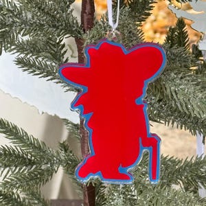 May include: A red and blue cowboy-shaped Christmas ornament. The ornament is hanging from a white ribbon and is displayed on a Christmas tree. The cowboy has a hat and a cane. The ornament is made of acrylic.