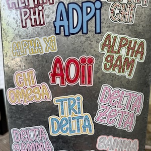 May include: A collection of colorful sorority and fraternity stickers, including "Alpha Phi," "ADPi," "Alpha Chi," and "Delta Zeta." The stickers feature various fonts and designs, with some incorporating floral patterns and glitter accents. They are displayed on a metallic surface.