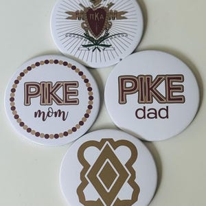 May include: Four white round buttons with brown and maroon designs. One button features a crest with the letters "ΠKA". Another says "PIKE mom" in a circle of dots. The third says "PIKE dad". The last has a geometric design.