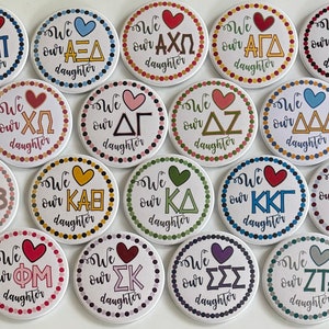 May include: A collection of 16 round buttons with white backgrounds and colourful borders. Each button features a different sorority Greek letter design with the text "We love our daughter" in black lettering.