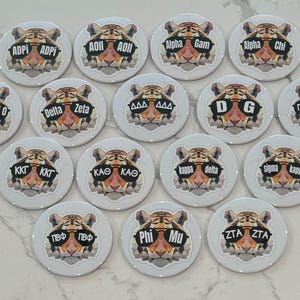 May include: A set of 15 round, white, tiger-themed buttons with different sorority names and initials. Each button features a cartoon tiger wearing sunglasses.