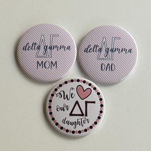 Puede incluir: Tres botones redondos con fondos a rayas rosa y blanco. Los dos botones superiores tienen el texto "delta gamma" y "MOM" en el botón izquierdo y "delta gamma" y "DAD" en el botón derecho. El botón inferior tiene un corazón rosa y el texto "We love our ΔΓ daughter" en un círculo negro con puntos rosa y negro.