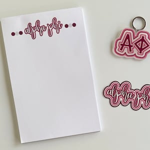 May include: A white notepad with the text "...alpha phi..." in maroon script with white dots on either side. A pink and maroon layered sticker with the text "alpha phi" and a pink and maroon layered keychain with the letters "AΦ" are also shown.