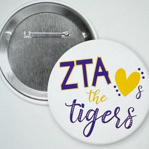 May include: A white button with a purple and gold design. The button says "ZTA the tigers" with a yellow heart.