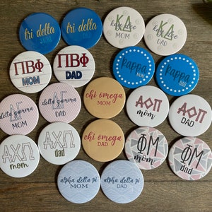 May include: A collection of 18 round buttons with various sorority and fraternity names and logos. The buttons are in different colors and patterns, including pink, blue, white, and gold. The text on the buttons includes "Tri Delta", "Kappa Alpha", "Phi Mu", "Alpha Delta Pi", "Chi Omega", "Delta Gamma", "Pi Beta Phi", and "Alpha Omicron Pi".