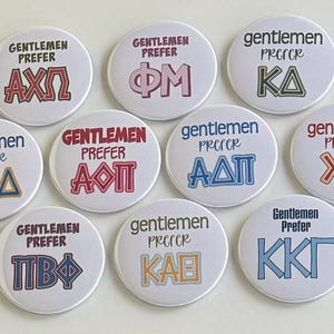 May include: Ten white round buttons with the text "Gentlemen Prefer" and different Greek letters in various colours. The letters include AXΩ, ΦΜ, KA, ΔΔΔ, ΑΟΠ, ΑΔΠ, ΧΩ, ΠΒΦ, ΚΑΘ, and ΚΚΓ.