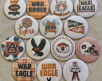 May include: A collection of fourteen circular pin-back buttons featuring various Auburn University themed graphics, including text like War Eagle, Let's Go Tigers, and War Damn Y'all, alongside illustrations of tigers, eagles, footballs, and helmets.
