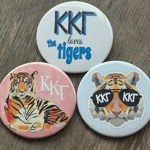 May include: Three circular badges with tiger designs. One badge has the text "KKΓ loves tigers" in blue. Another features a tiger on a pink background with "KKΓ" in white. The third badge shows a geometric tiger with sunglasses.