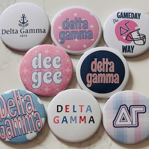 May include: Eight circular pin-back buttons featuring Delta Gamma sorority designs. The buttons display various patterns, including pink stars, blue polka dots, plaid, and solid colors, with text such as Delta Gamma, dee gee, and The Gameday Way.