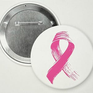 Può includere: Un bottone bianco con un design a nastro rosa, simbolo della sensibilizzazione al cancro al seno. Il nastro è dipinto con pennellate. Il retro del bottone è argentato con una spilla.