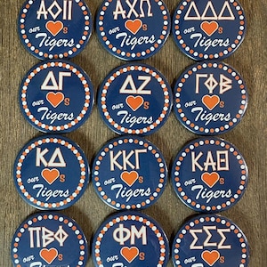 Puede incluir: Doce botones circulares azules y blancos con lunares blancos alrededor del borde. Cada botón presenta un nombre de hermandad de letras griegas diferente en blanco con un corazón rojo y el texto "our Tigers" en blanco.