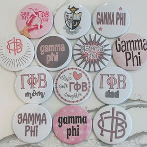 Peut inclure: Une collection de 12 boutons ronds avec différents motifs. Les boutons présentent les lettres grecques Gamma Phi Beta, les mots "Gamma Phi" et d'autres motifs. Les boutons sont blancs avec des accents roses et bruns.