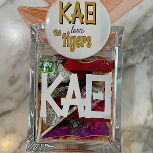 May include: A clear glass jar filled with colorful candies and a white sticker with the letters "KAΘ" on it. A round button with the text "KAΘ the loves tigers" is on top of the jar.